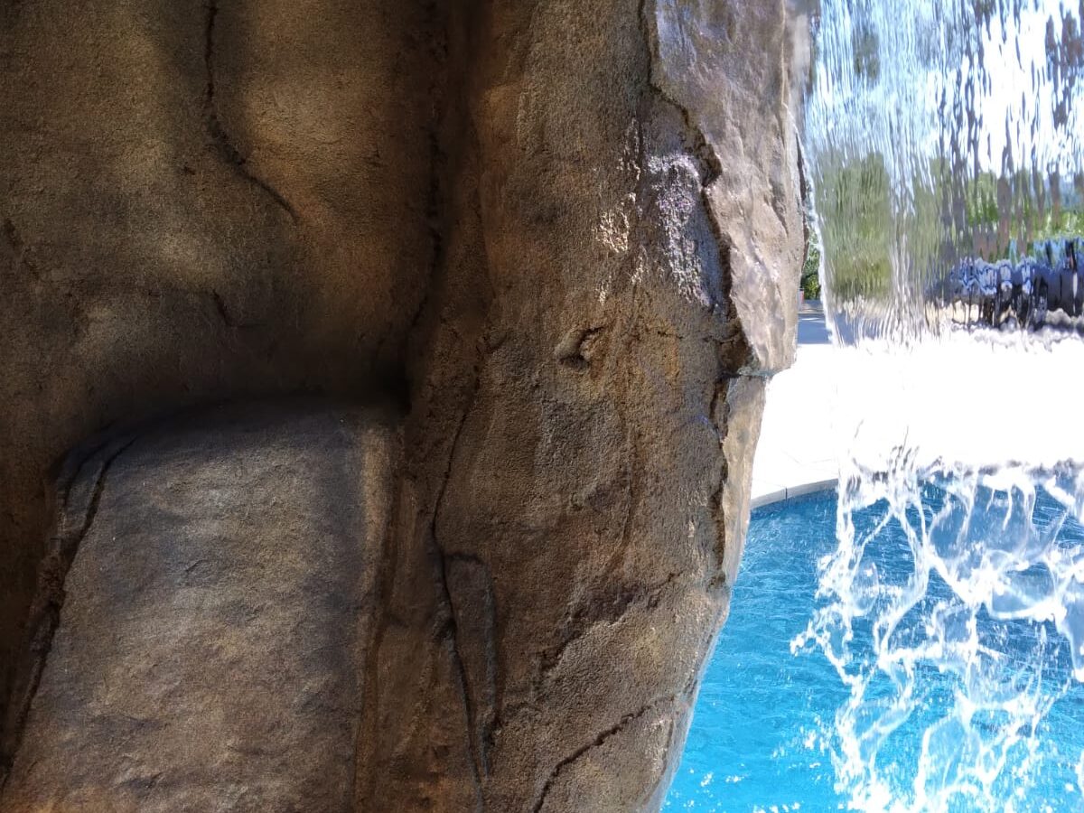 View from inside an artificial rock grotto looking through a curtain waterfall to a bright blue pool